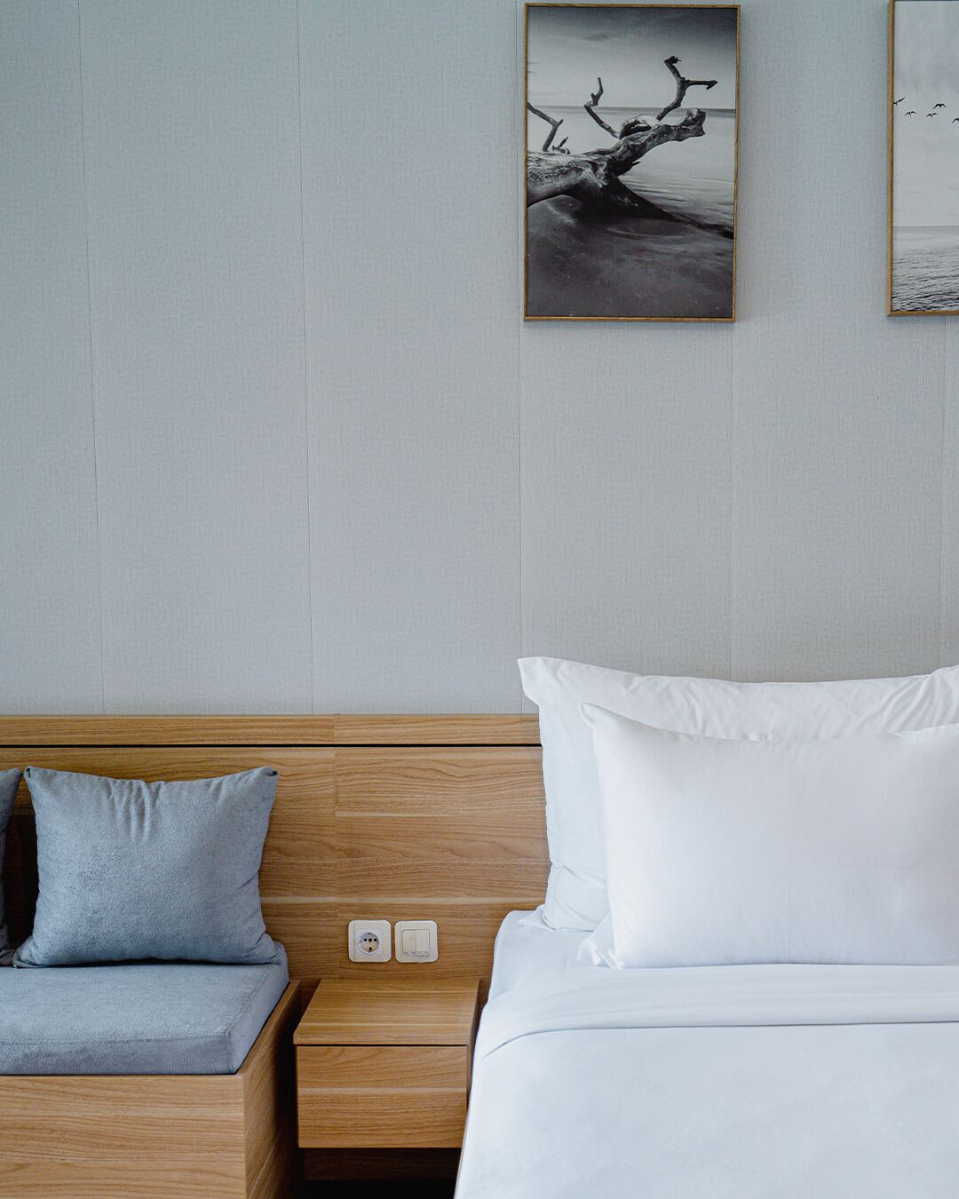 A hotel room with a wooden headboard, a bed with white bedding, a grey pillow, a nightstand with a phone and a power outlet, and two framed pictures on the wall.