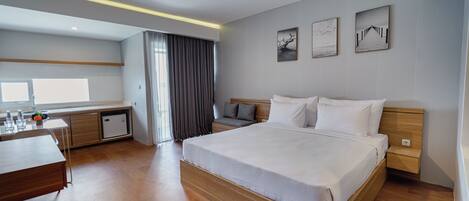 A modern hotel room with a large bed, a desk, a small kitchenette, and a view of the outdoors.