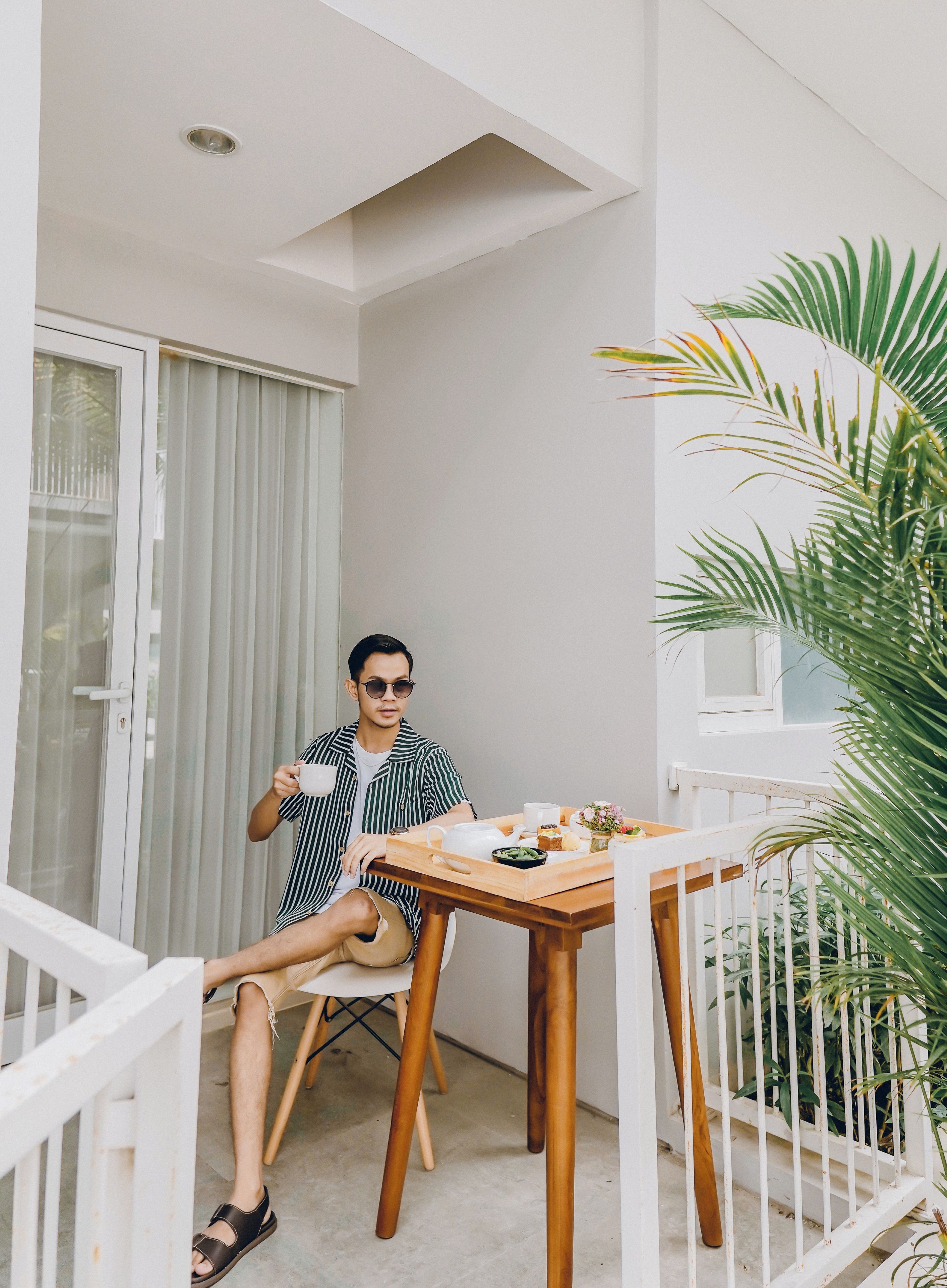 A man sitting at a wooden table on a balcony with a white railing, holding a cup. There is a potted plant to the right and a window with blinds in the background.
