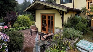 Een huis met twee verdiepingen, voorzien van een overdekt terras, een houten terras en een tuin met verschillende planten en bloemen.