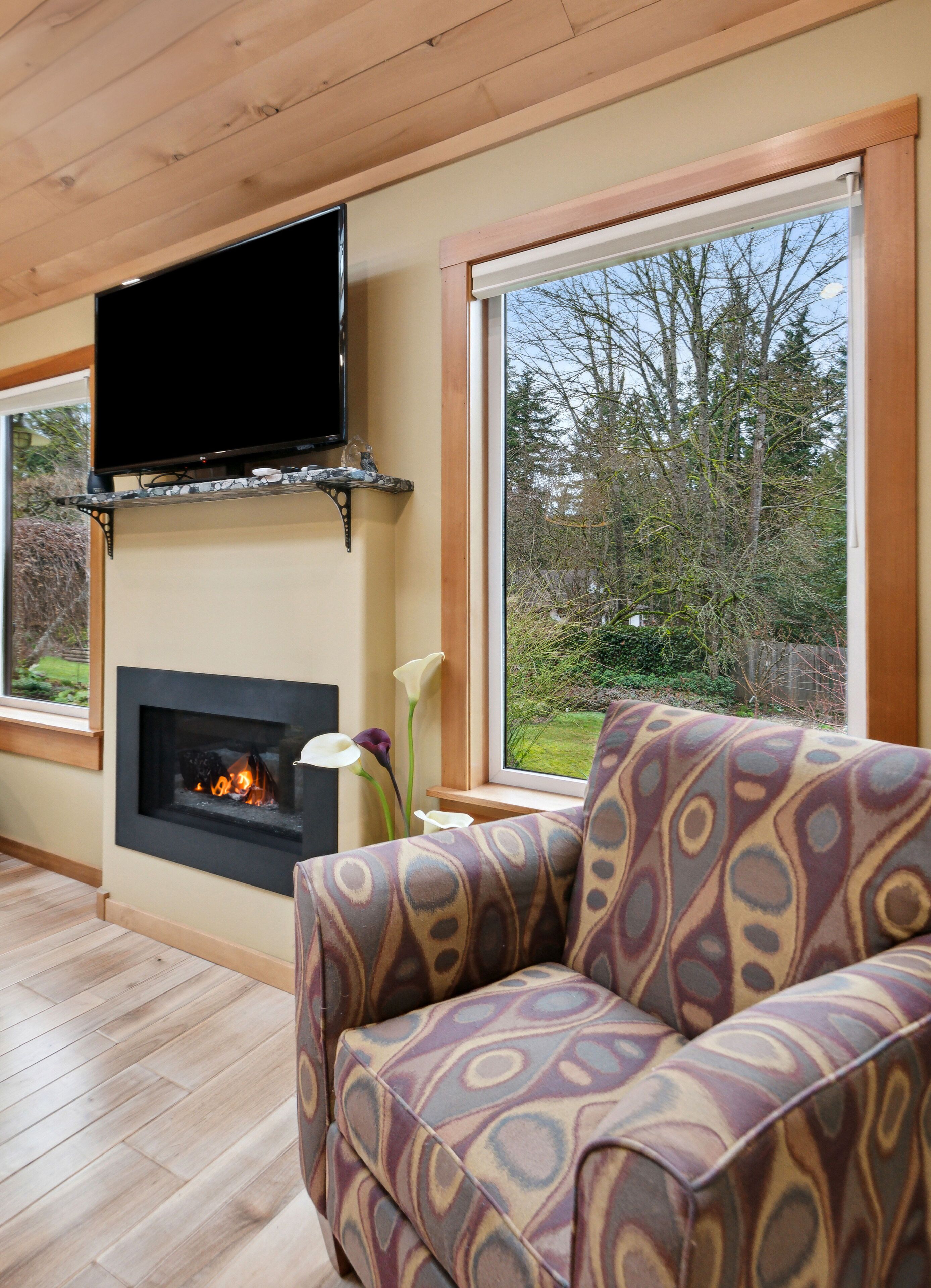 A cozy living room with a fireplace, a flat-screen TV mounted above it, and a large window offering a view of the outdoors.