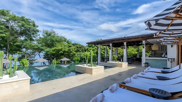 A poolside area with lounge chairs, a pool, and a covered seating area with umbrellas. - Beach Club Included | 1 min to beach | sleeps 5 | Plaza Mercado 12 (Las Catalinas)