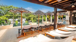 A poolside area with sun loungers, striped umbrellas, and a view of a pool and greenery. - Beach Club Included | 1 min to beach | sleeps 5 | Plaza Mercado 12 (Las Catalinas)