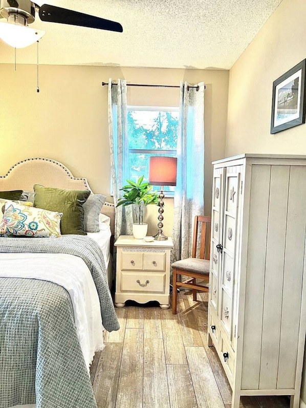 Un dormitorio con cama, mesita de noche, lámpara, silla, armario y ventana con cortinas.