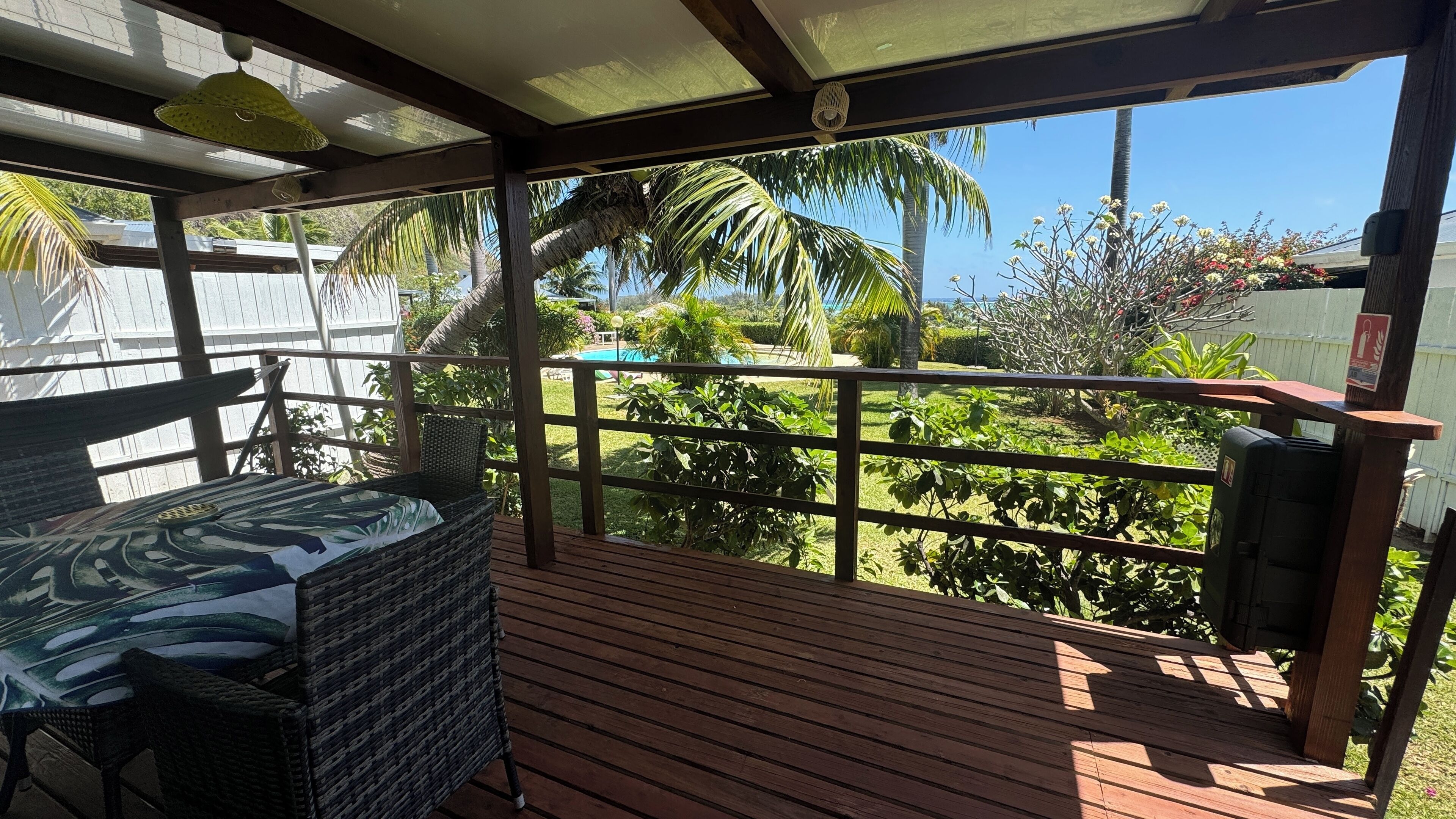 Un espace extérieur abrité, doté d’une terrasse en bois, d’un fauteuil en osier et offrant une vue sur un jardin avec des palmiers et une piscine.