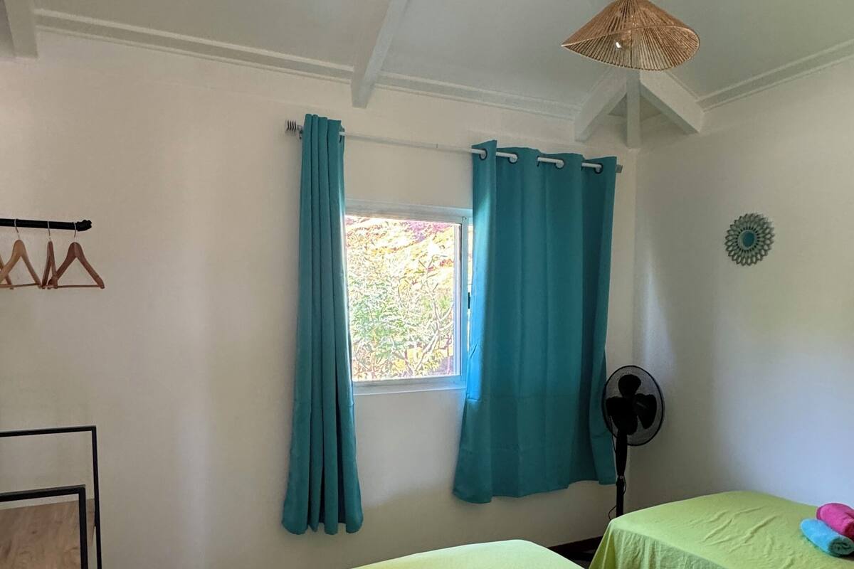A room with two beds, a window with curtains, a fan, and a ceiling fan.