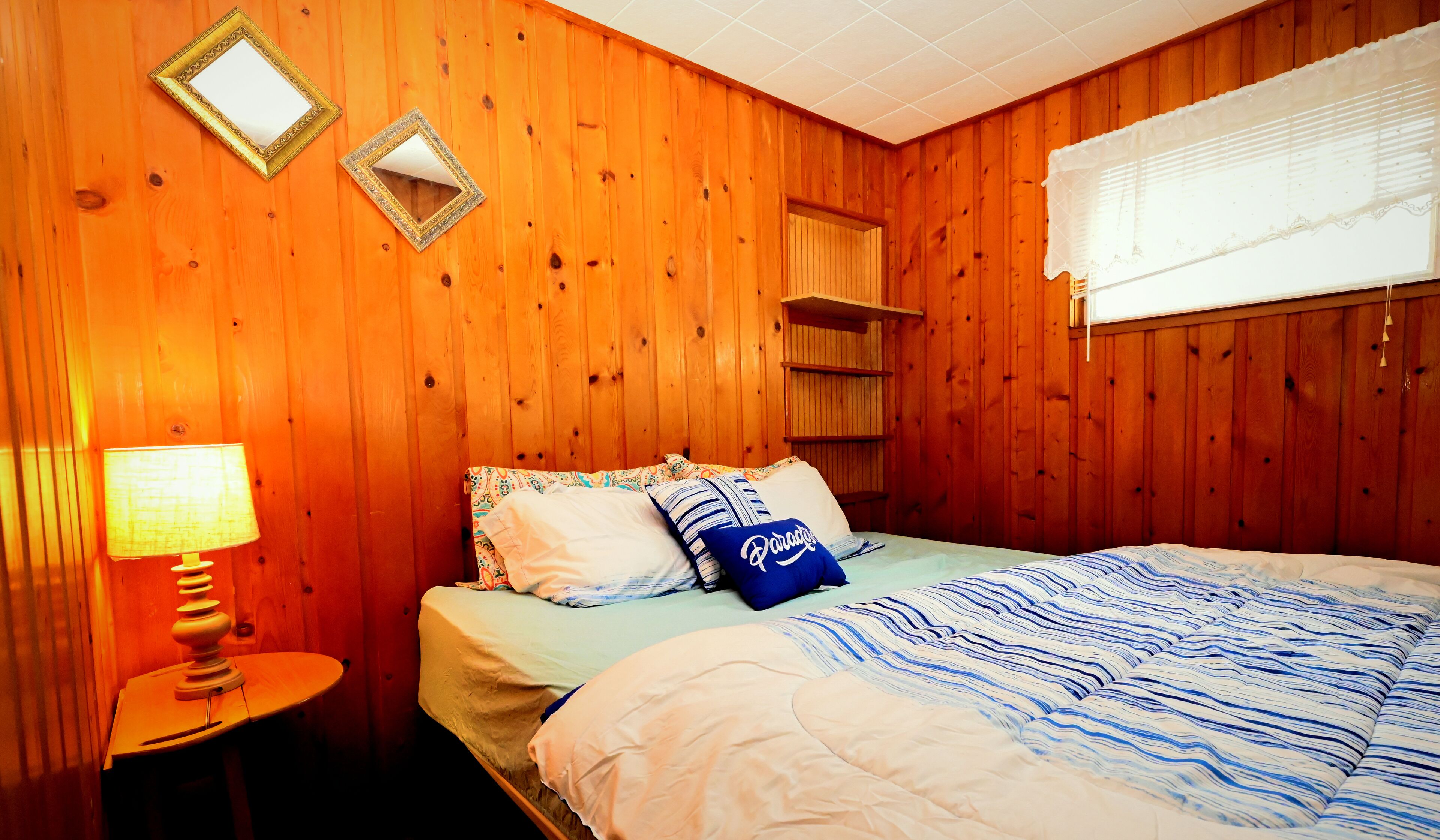 A wooden-paneled room with a bed, a bedside table with a lamp, and two wall mirrors.