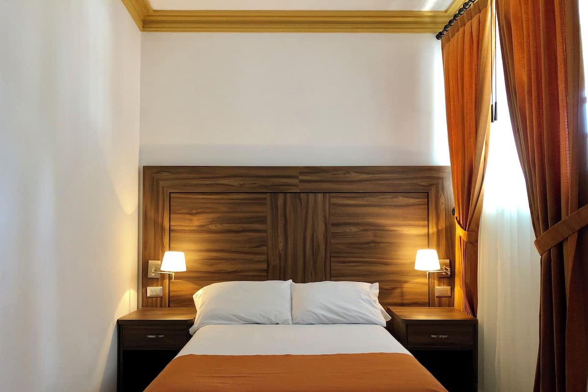 Habitación de hotel con cabecera de madera, dos lámparas de noche, cama con ropa blanca y cortinas marrones.