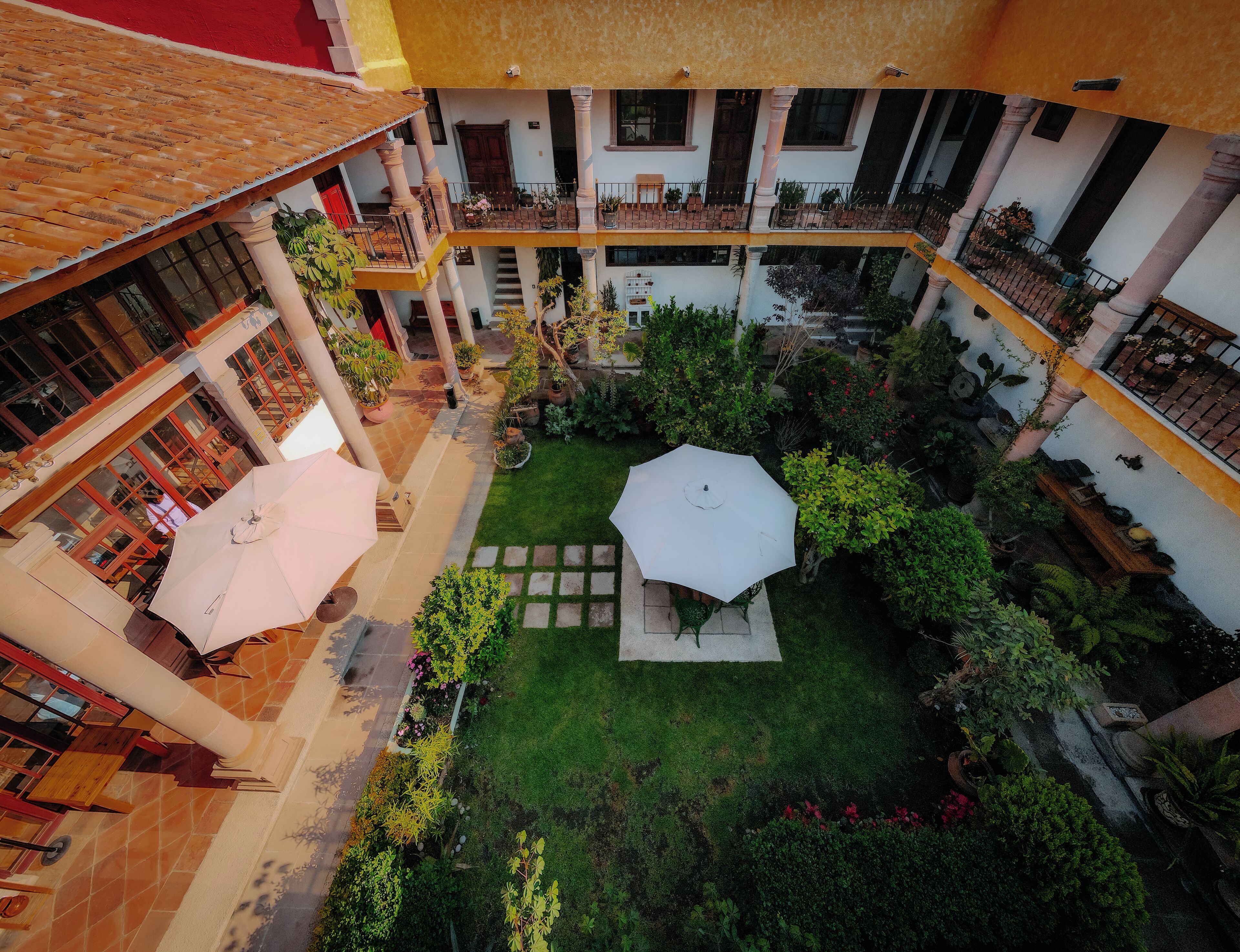 Une cour intérieure avec une table octogonale au centre, entourée de verdure, et bordée de balcons à plusieurs niveaux, dotés de rampes et de plantes en pot.
