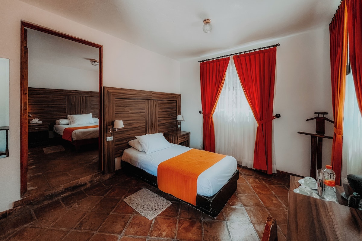 Habitación de hotel con cabecera de madera, cama con ropa blanca y una manta naranja, mesita de noche con lámpara, espejo y cortina roja.