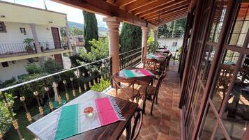 Un espacio exterior cubierto para comer con mesas y sillas de madera, piso de baldosas y vistas de los edificios y espacios verdes circundantes.
