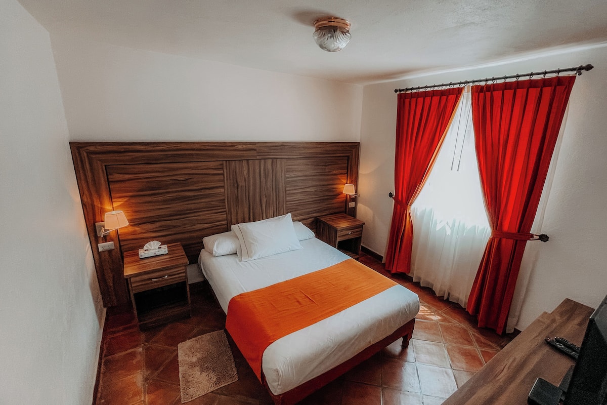 Habitación de hotel con cama de cabecera de madera, dos mesitas de noche con lámparas, cortina roja y suelo de baldosas.