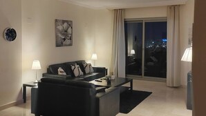 A modern living room with a black sofa, two side tables with lamps, a coffee table, a large window with curtains, and a cityscape view at night. - 2 Bed Flat-Sleeps4-Pets-Pool-Parking-GolfClub (Estepona)