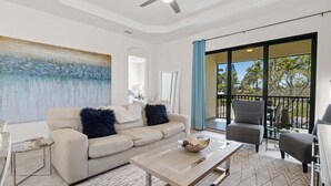 Ein modernes Wohnzimmer mit einem beigen Sofa, einem Couchtisch und einem grauen Sessel. Ein großes Fenster bietet Blick auf Bäume, und ein Balkon lädt mit einem Stuhl zum Verweilen ein.