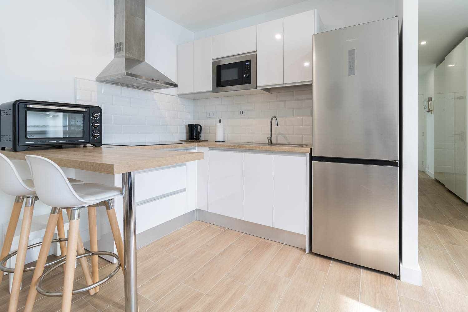 A modern kitchen with white cabinets, a stainless steel refrigerator, a microwave, and a toaster. There are two white bar stools at the counter.
