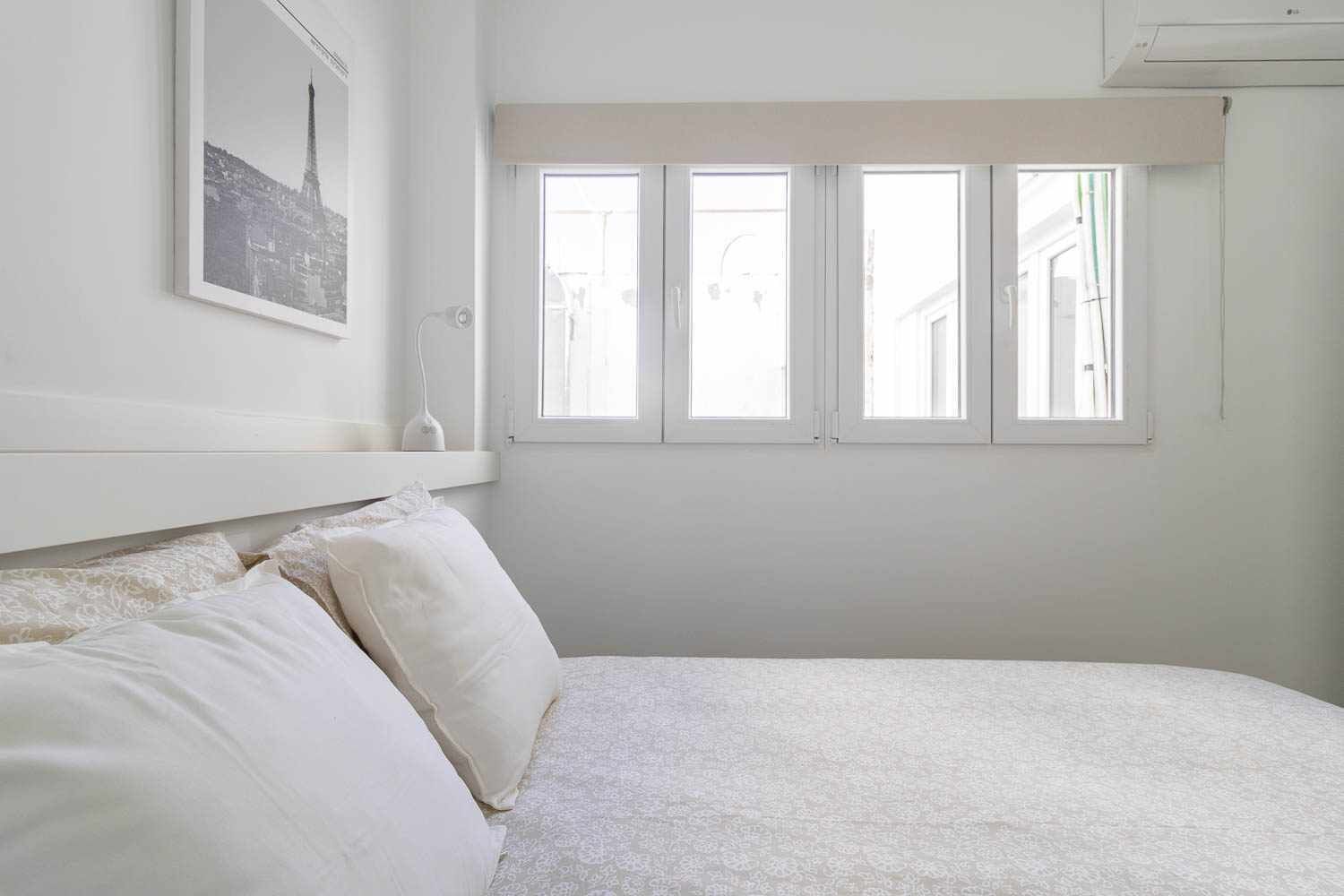 Un dormitorio con cama, ropa de cama blanca, un cuadro enmarcado en la pared y una ventana con vistas a edificios.