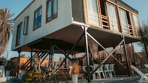 A two-story building with a balcony, supported by metal beams, surrounded by various plants and outdoor furniture.