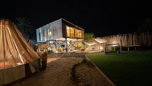 A modern building with a large window and a balcony, surrounded by a sandy area and a grassy field.
