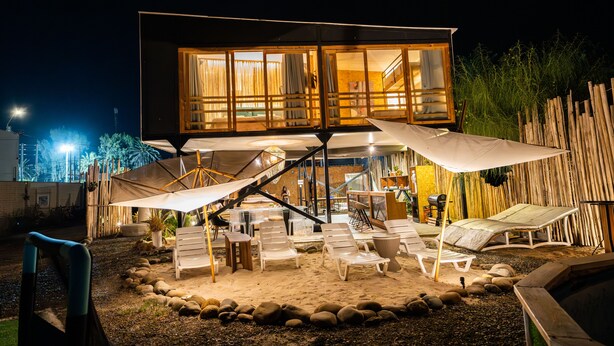 A modern outdoor lounge area with multiple seating options, a staircase leading to an upper level, and a well-lit interior visible through large windows.