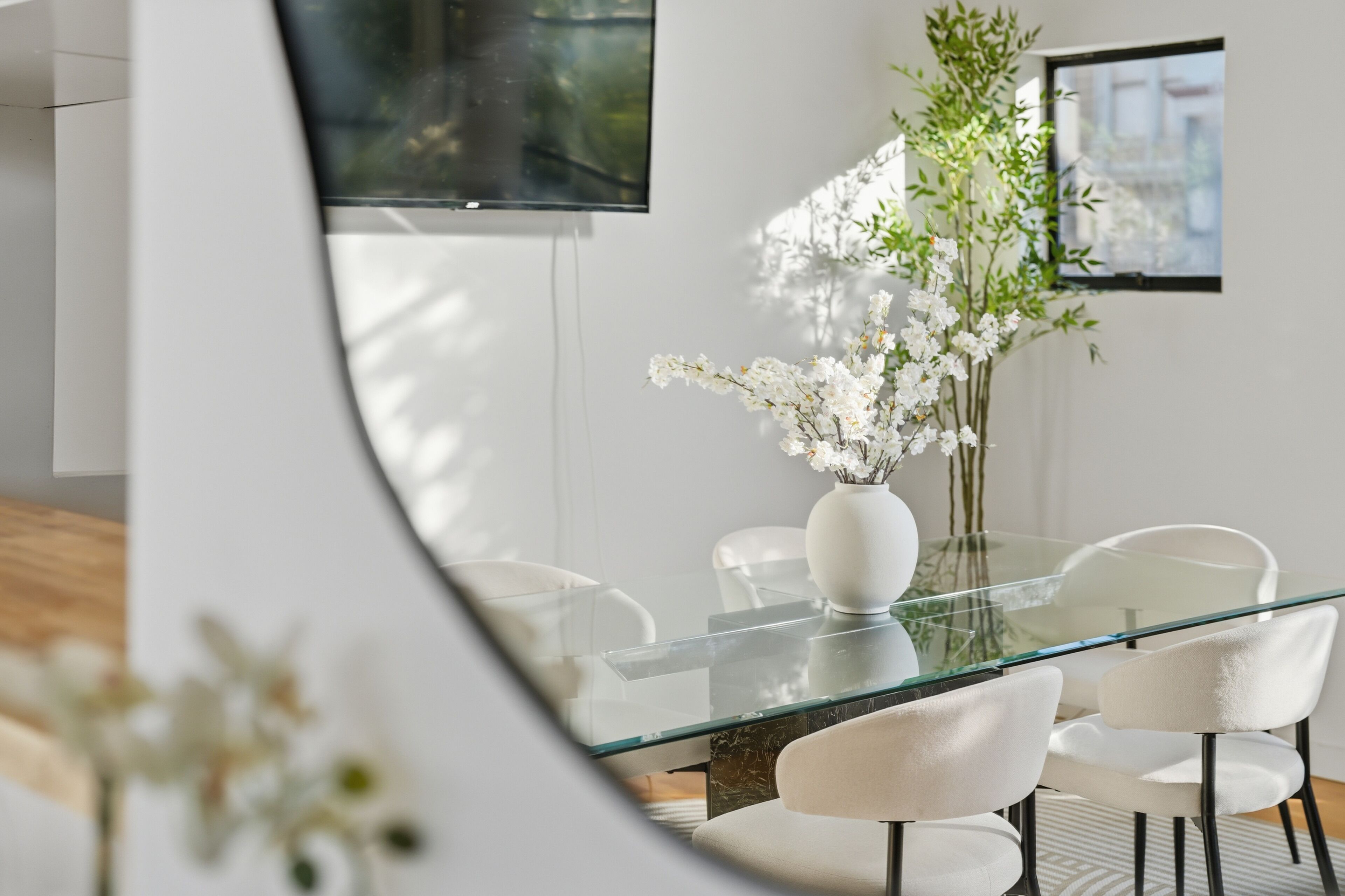A modern dining area with a glass table, white chairs, and a vase of white flowers.