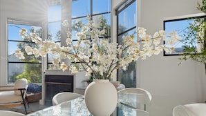 En modern inredning med ett glasbord, vita stolar, en vas med vita blommor och stora fönster.