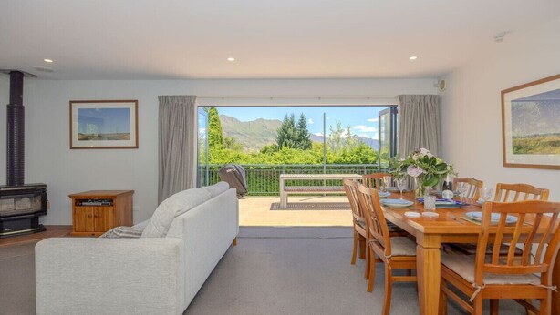 A living room with a sofa, a dining table set for four, and a view of mountains through large windows. - Terrific views on Totara (Wānaka)