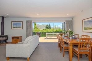 A living room with a sofa, a dining table set for four, and a view of mountains through large windows. - Terrific views on Totara (Wānaka)