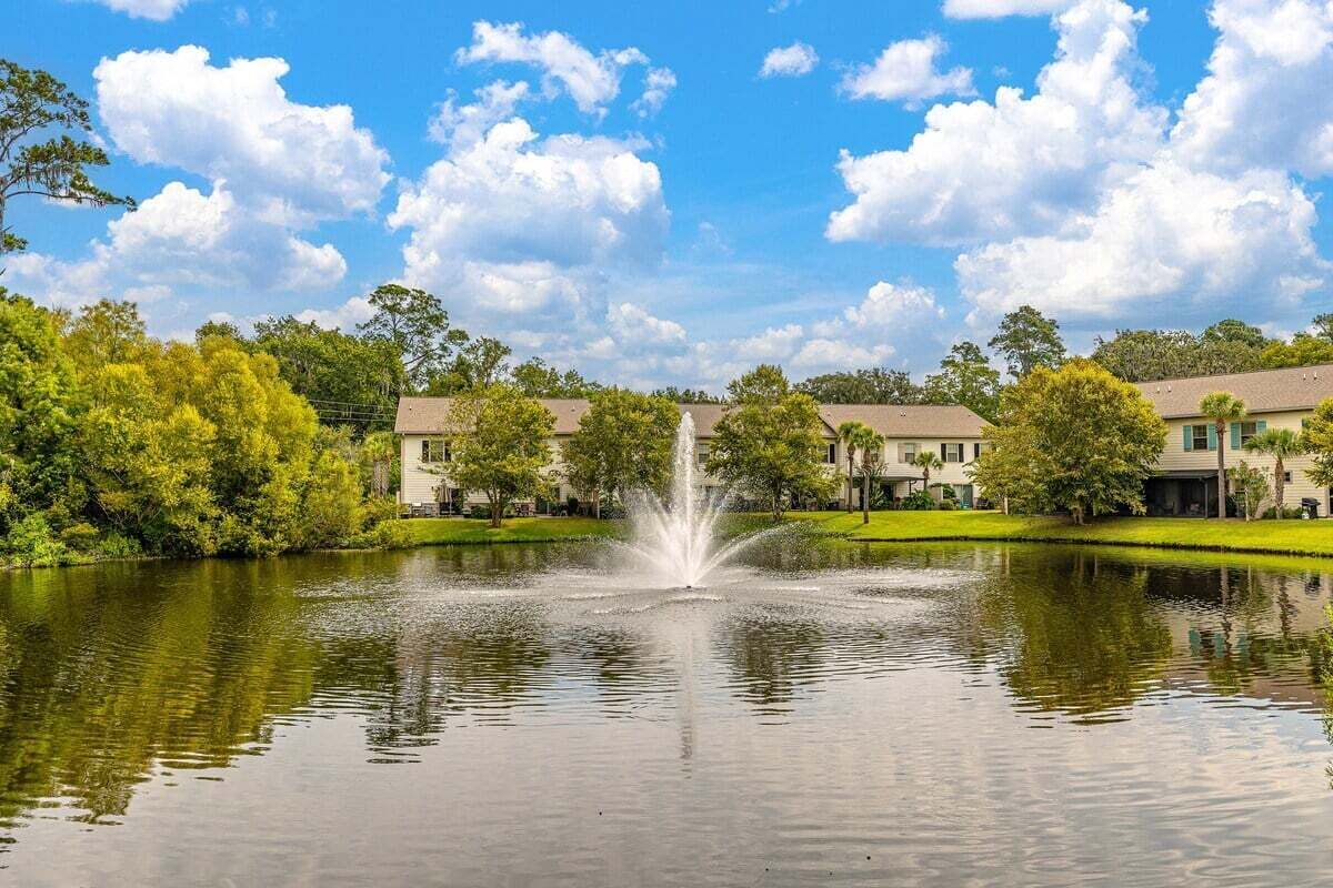 A serene pond with a fountain in the center, surrounded by lush greenery and a building with a flat roof in the background.