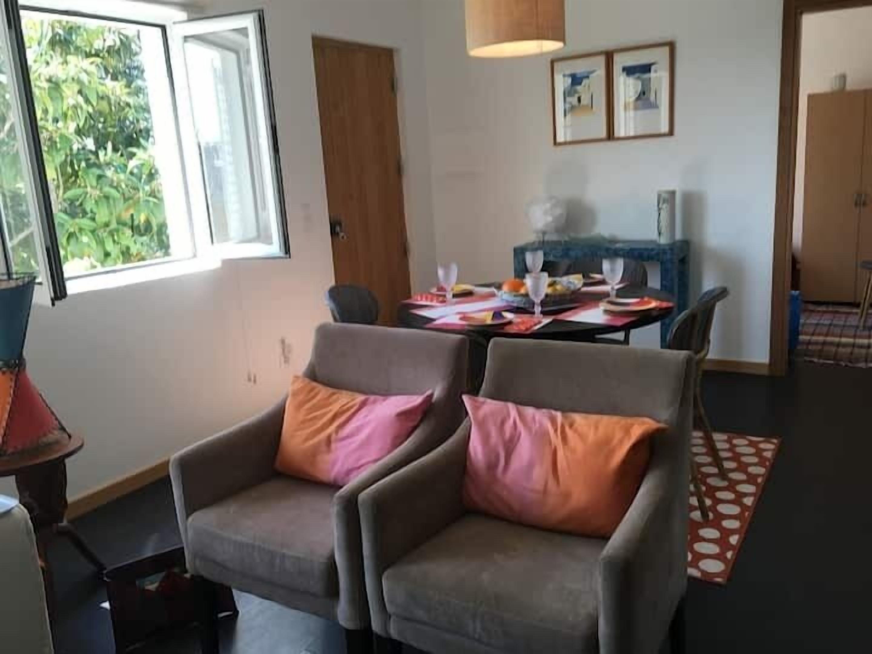 A room with two grey armchairs, a dining table set with plates, glasses, and a vase of flowers. There is a window with a view of greenery, a door, and a wall with two framed pictures.