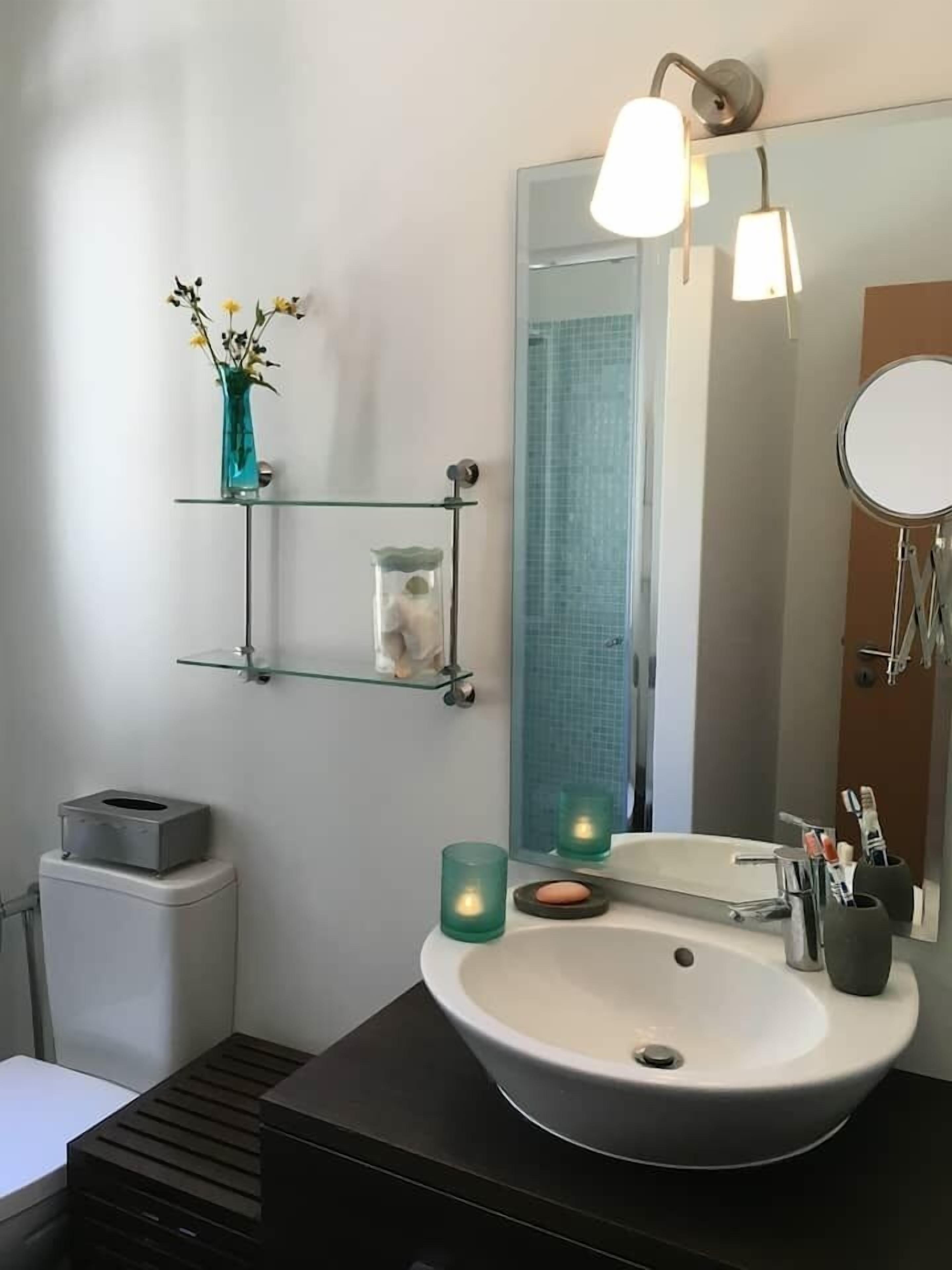 Une salle de bain moderne avec un grand miroir, un lavabo blanc et une étagère en verre où sont rangés les articles de toilette.