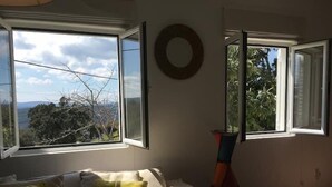 A room with a bed, a window with an open shutter, and a view of trees and hills.