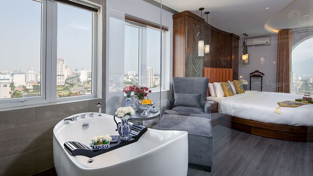 A modern hotel room with a large window, a bathtub, a grey sofa, a wooden headboard, and a large bed. - TGT BEACH HOTEL (Da Nang)