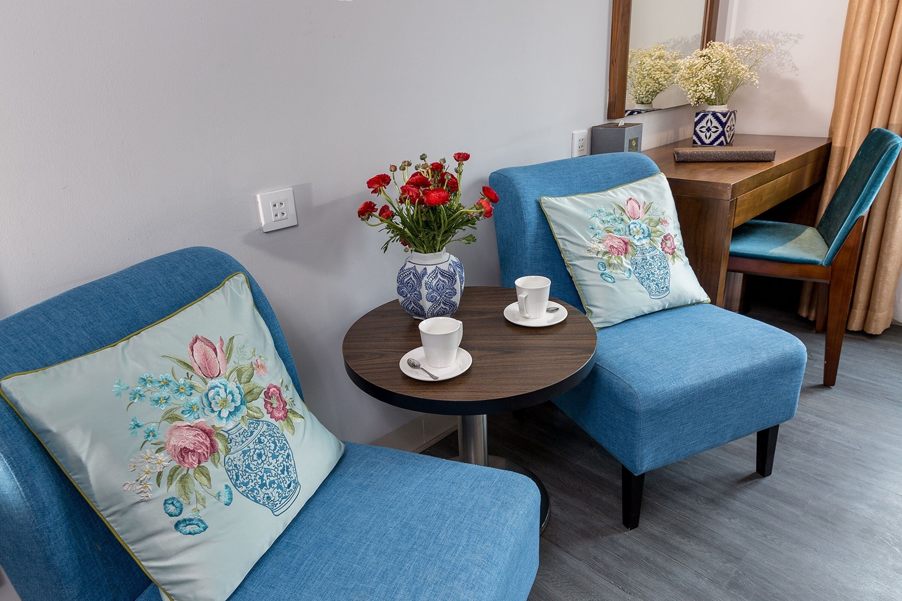 A room with two blue armchairs, a small round table with a vase of red flowers, and a mirror above the table.