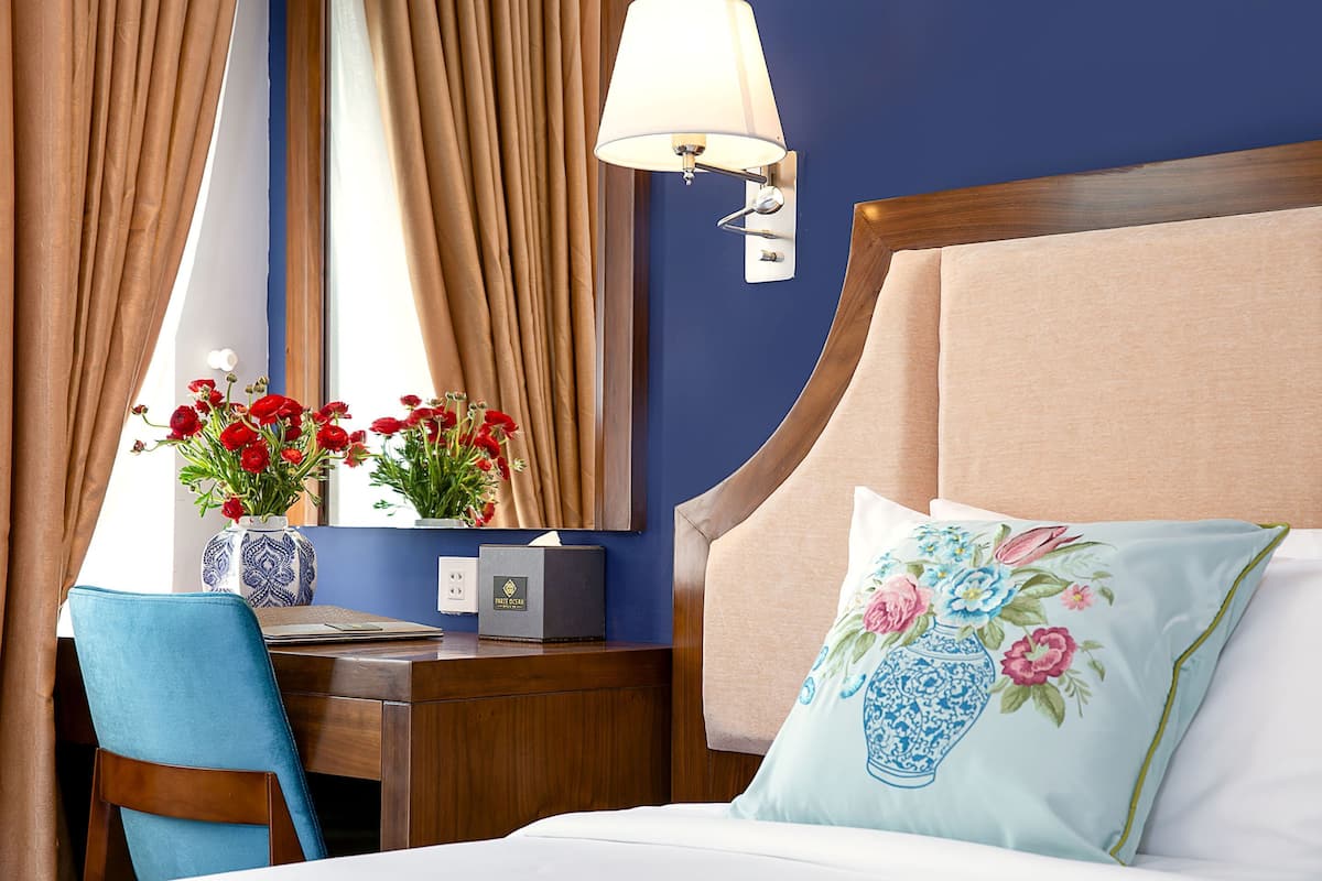 A hotel room with a bed, a desk, a chair, a vase with flowers, and a lamp.
