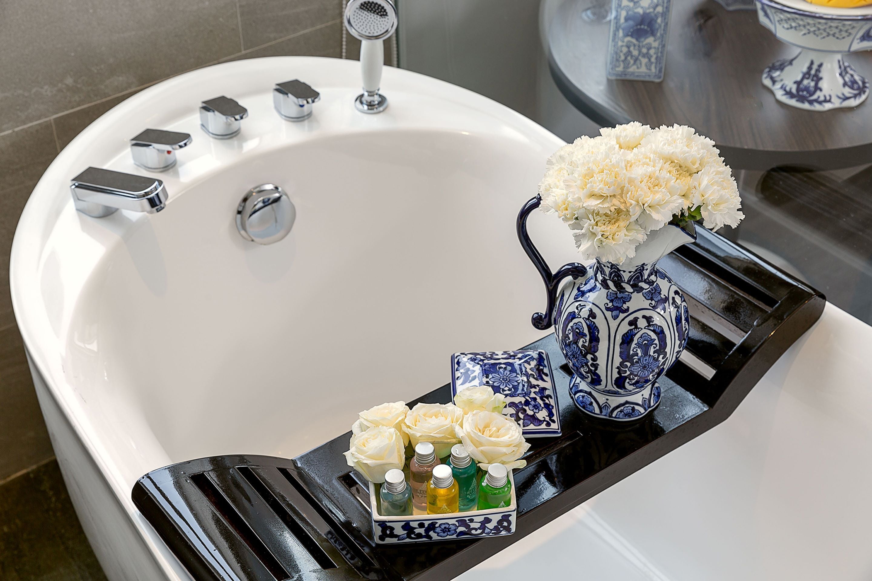A bathroom with a white bathtub, a black tray with toiletries, and a blue and white vase with white flowers.