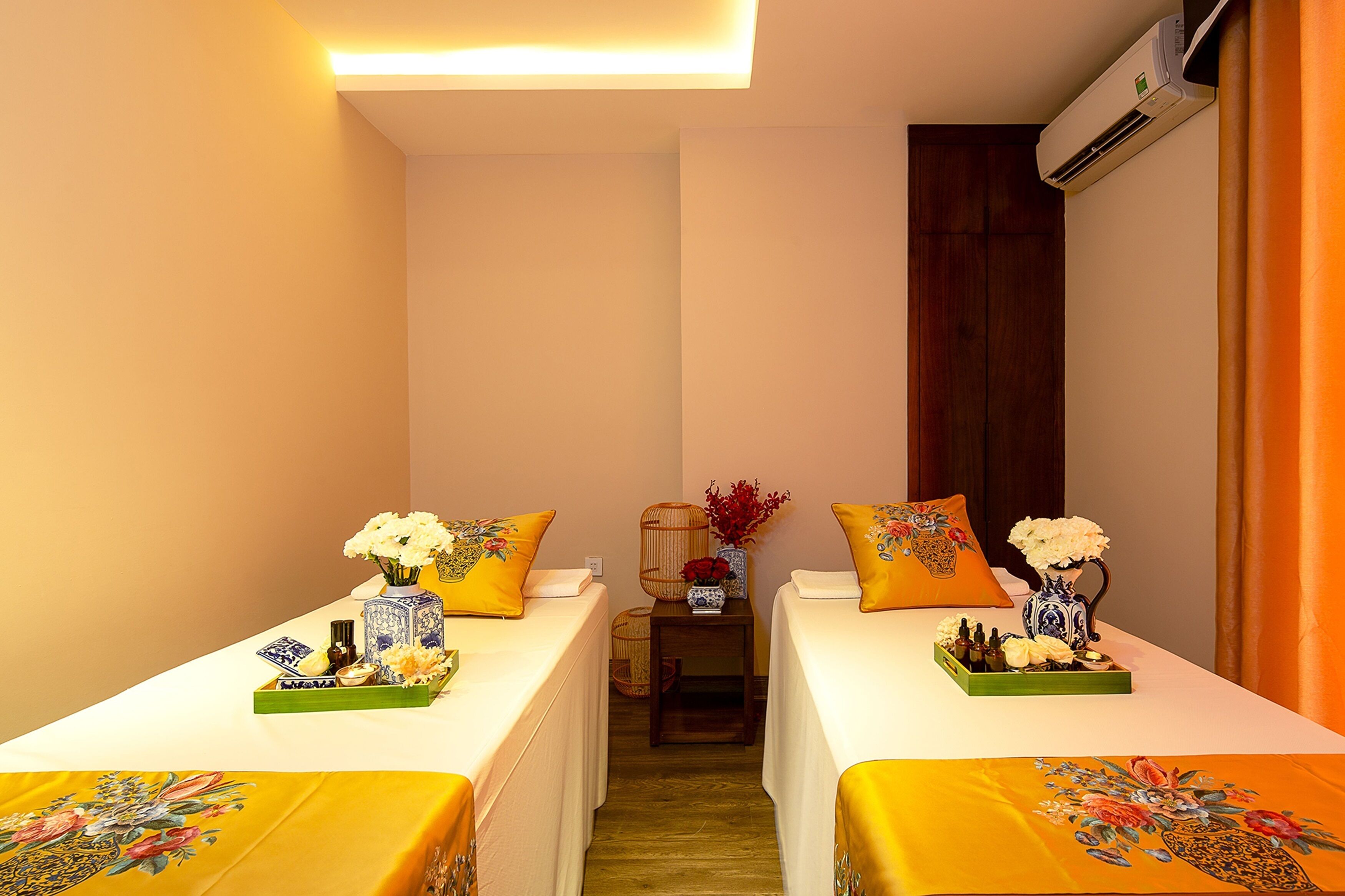 Habitación de hotel con dos camas, cada una con cubrecama y almohadas amarillas a juego. Hay jarrones decorativos con flores sobre las camas, una mesita con una bandeja de objetos y un equipo de aire acondicionado en la pared.