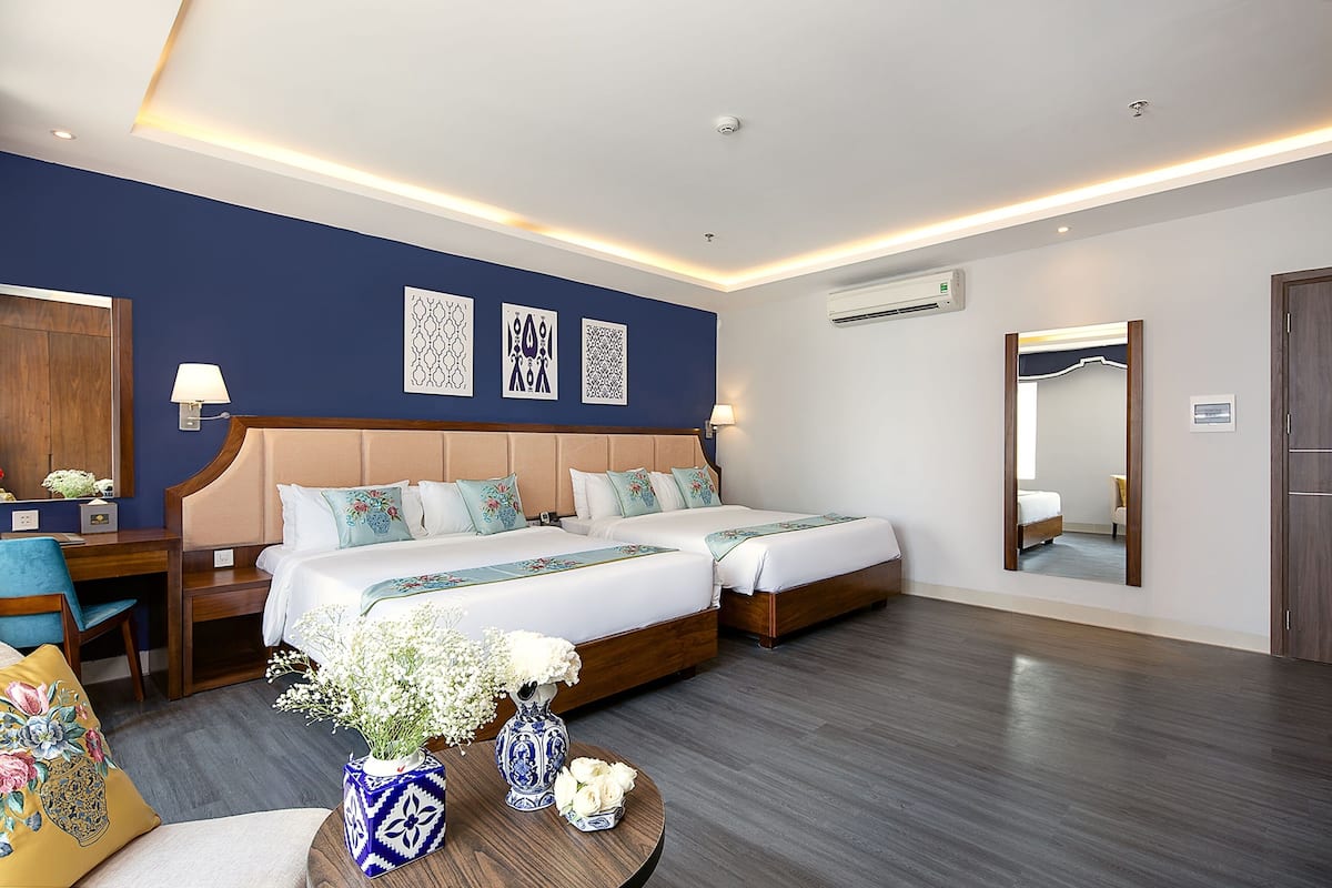 A hotel room with two beds, a desk, a chair, a round table with vases, and decorative wall art.
