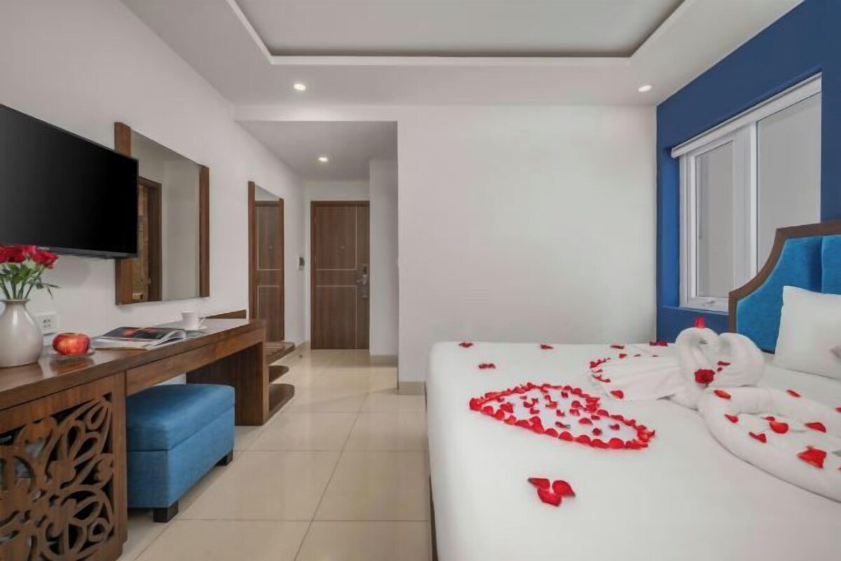 A hotel room with a bed adorned with rose petals and swans, a desk with a flat-screen TV, a blue chair, and a wooden cabinet.
