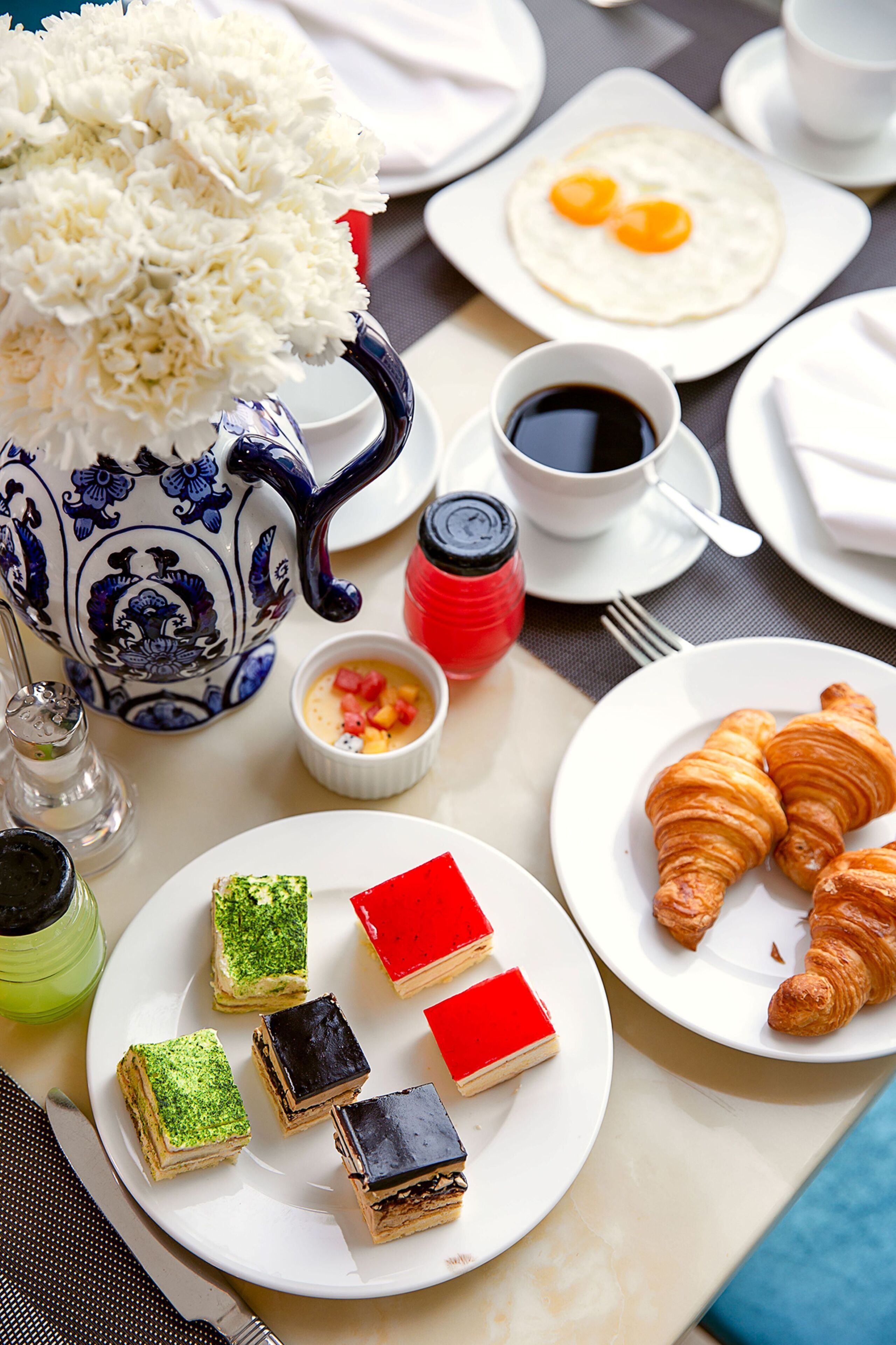 Una mesa dispuesta con diversos alimentos para el desayuno, incluyendo pasteles, un croissant, un plato de huevos y un pequeño jarrón con flores.