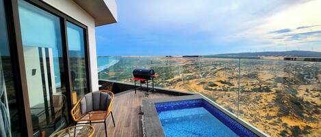 A balcony with a pool, wicker chairs, and a grill overlooking a desert landscape.