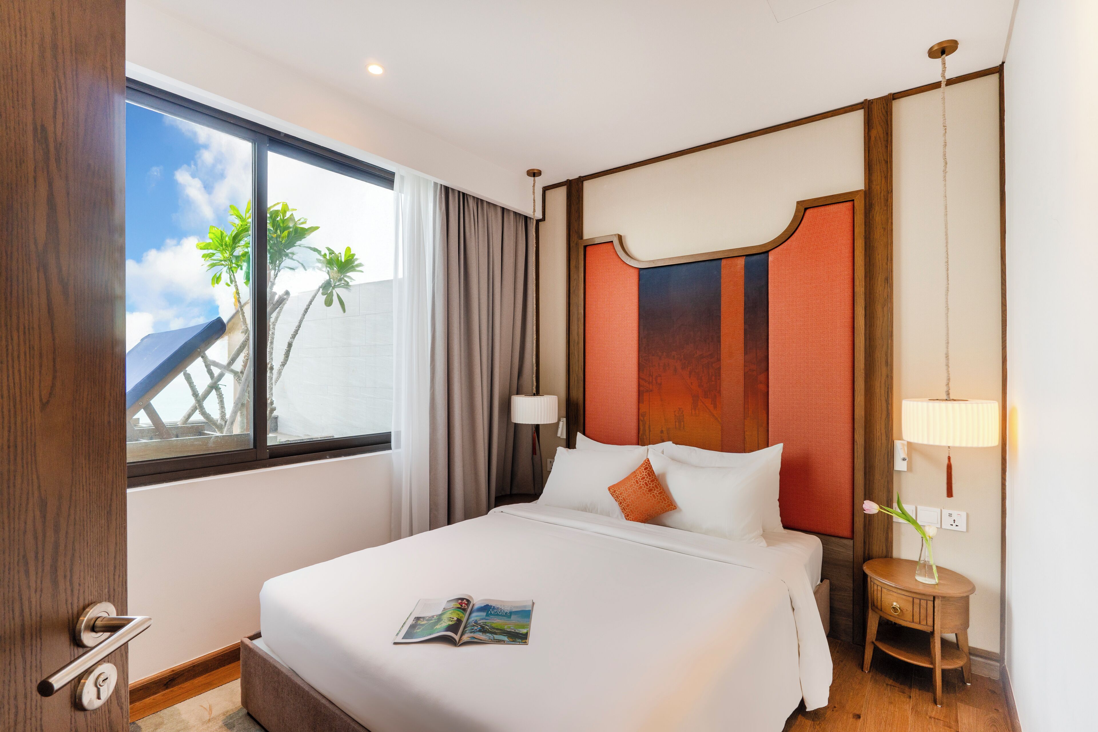A hotel room with a large bed, a bedside table with a lamp, a window with a view of palm trees, and a door with a key lock.