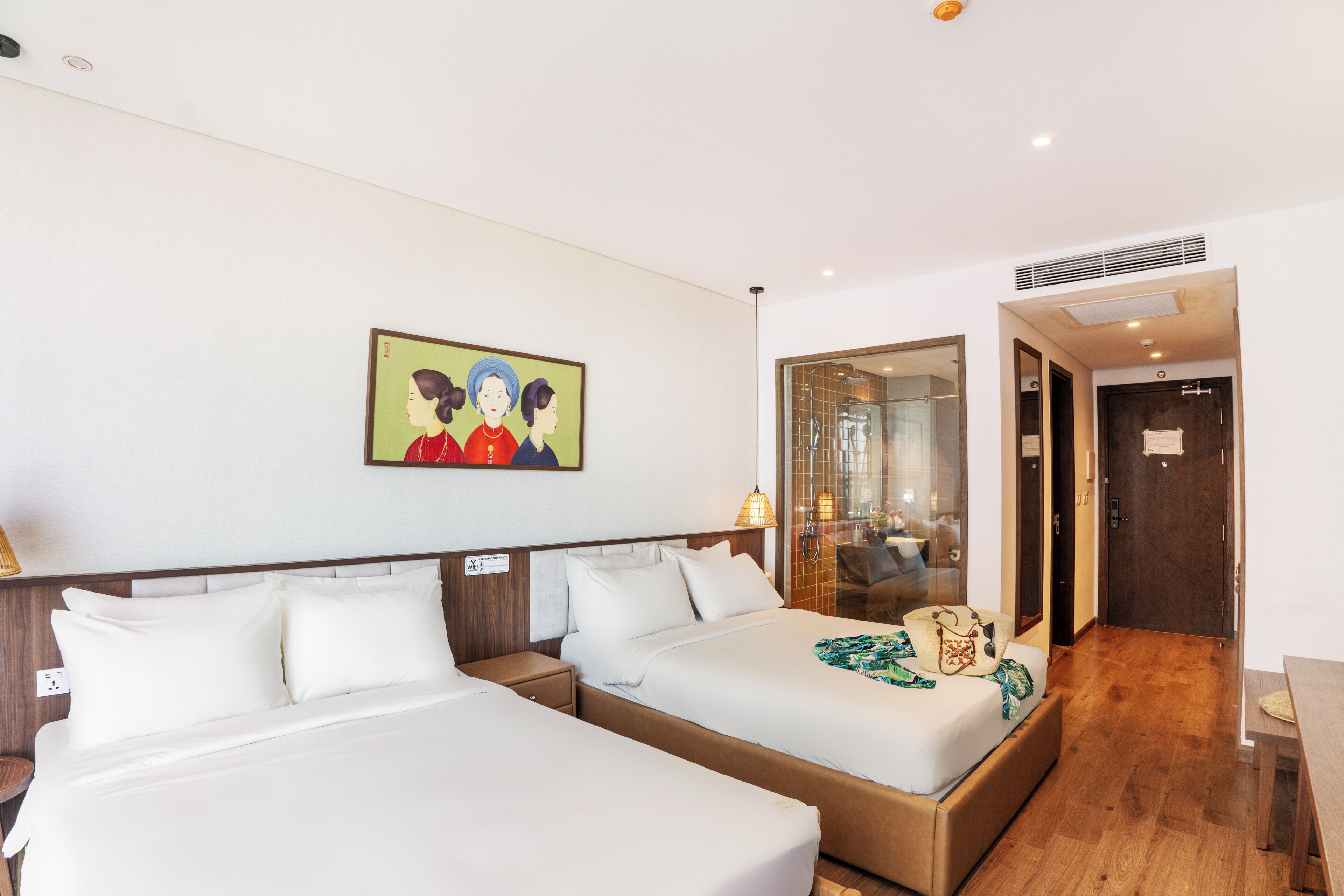 A hotel room with two beds, a painting on the wall, and a bathroom visible through a glass door.