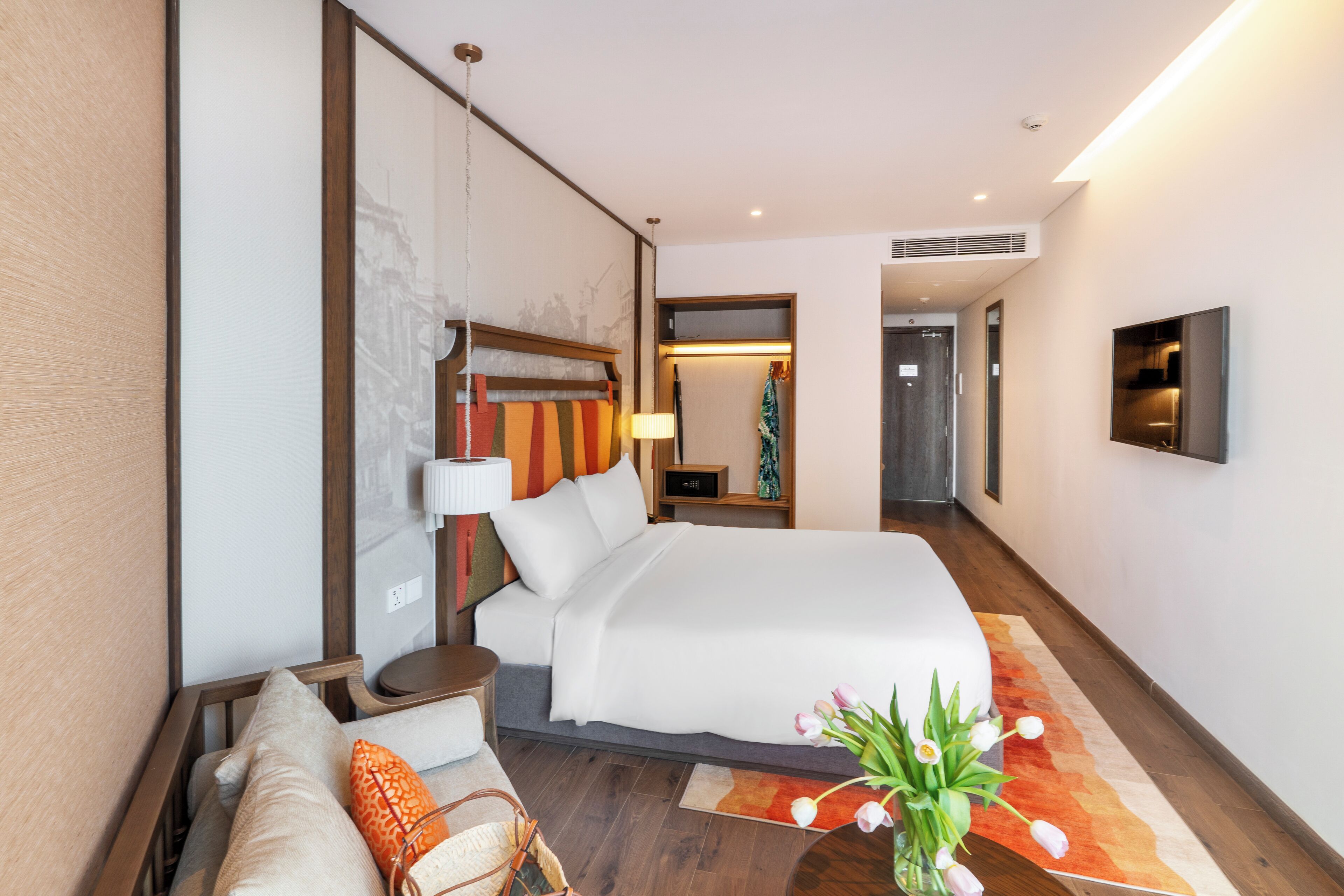 A modern hotel room with a large bed, a sofa, a nightstand, a lamp, and a TV mounted on the wall.