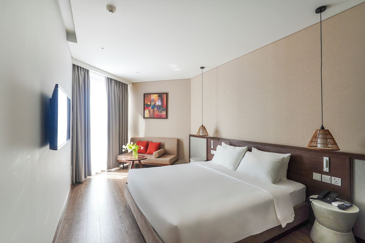 A hotel room with a large bed, a sofa, a small table with a vase, and a painting on the wall.