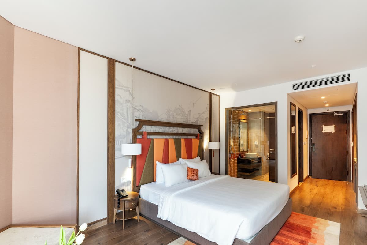 A modern hotel room with a large bed, a wooden headboard, and a view of the bathroom through a glass door.