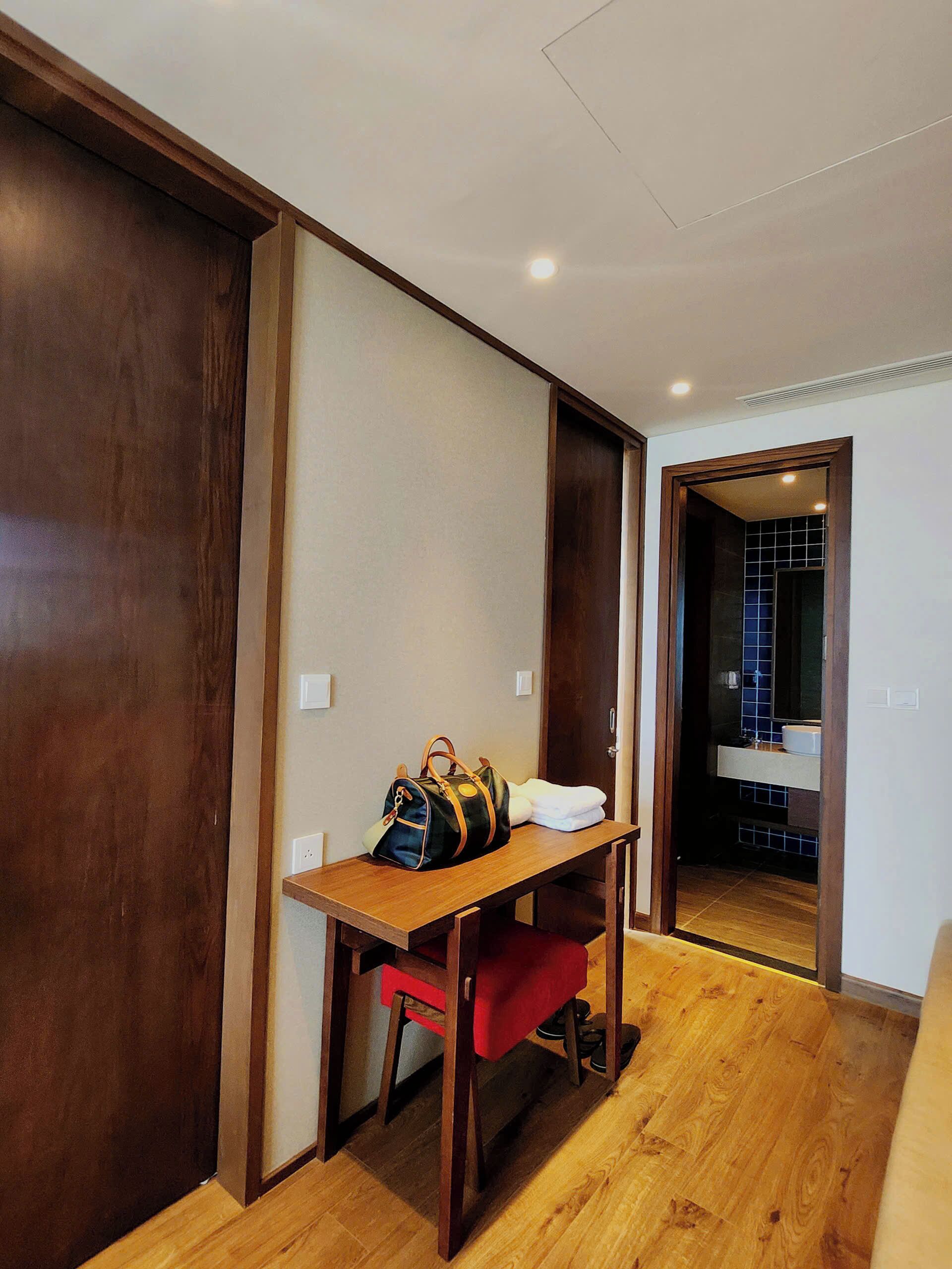Habitación de hotel con un escritorio de madera, una silla roja, un espejo grande y un baño visible a través de una puerta abierta.