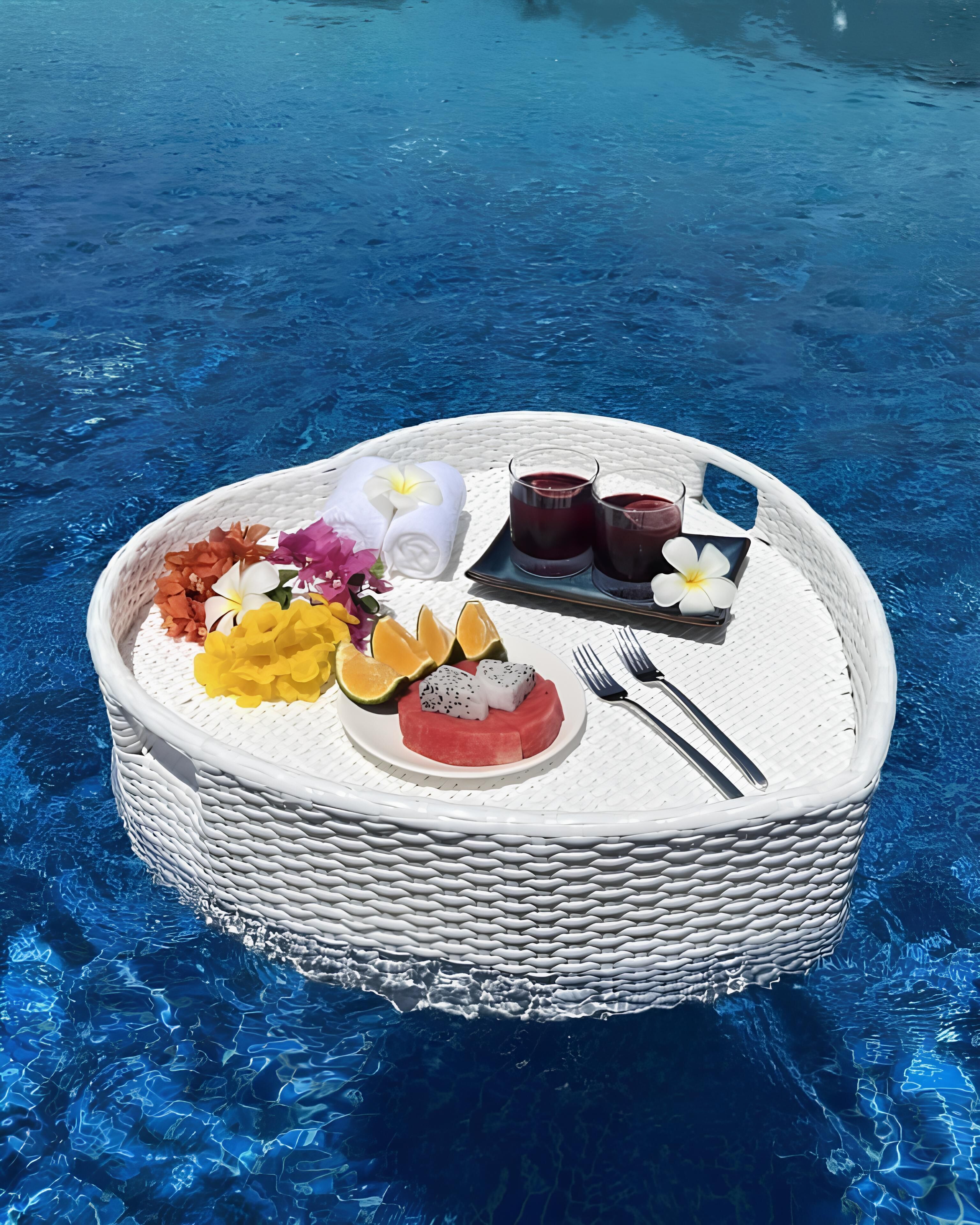 Une table ronde en osier, recouverte d’une nappe blanche, est placée au bord d’une piscine dont l’eau est d’un bleu limpide.