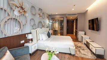 Quarto de hotel moderno com uma cama grande, uma mesa de cabeceira, uma cadeira, uma mesinha com um vaso e uma TV fixada na parede.