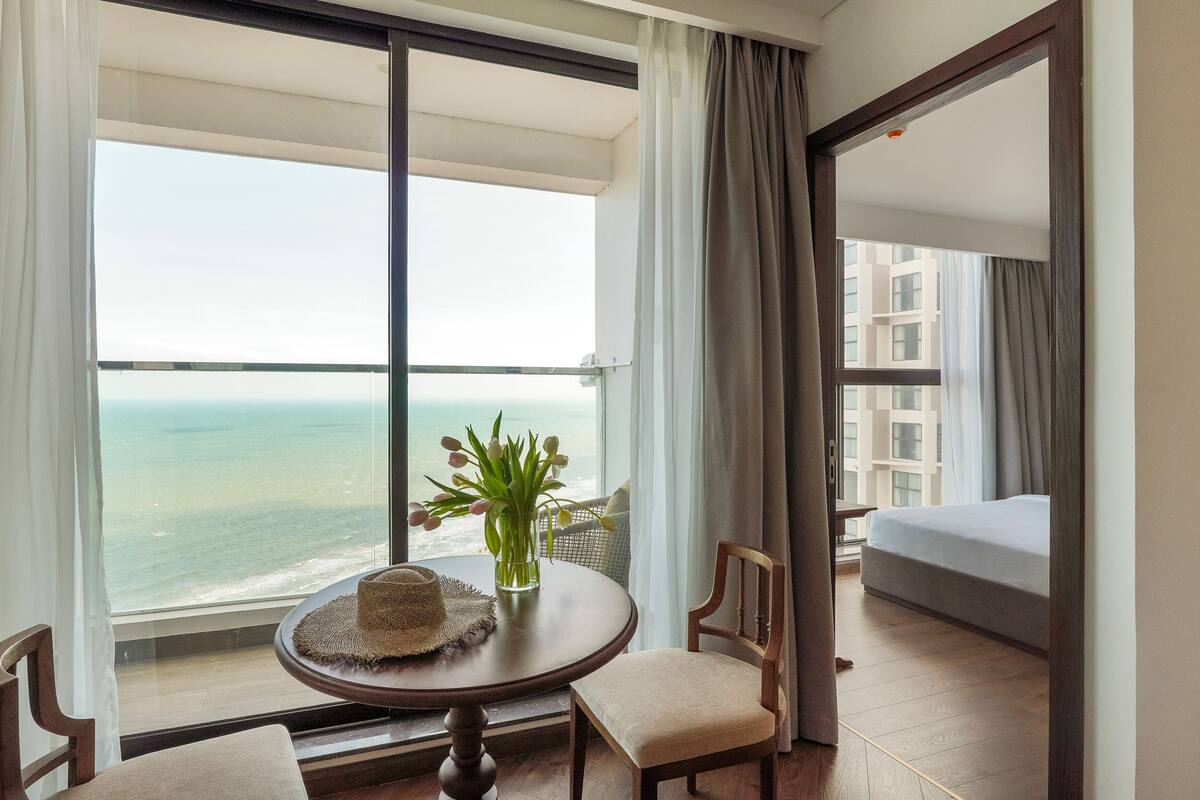 A hotel room with a large window, a round table with a hat, a chair, a small plant, and a bed.