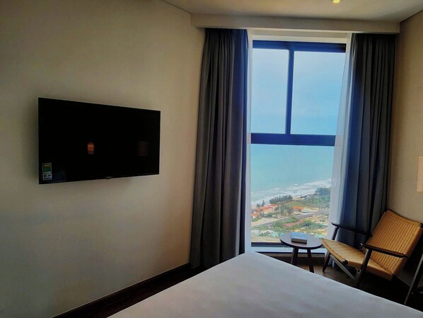 A hotel room with a large bed, a flat-screen TV mounted on the wall, a chair, and a view of a coastal landscape through a large window.